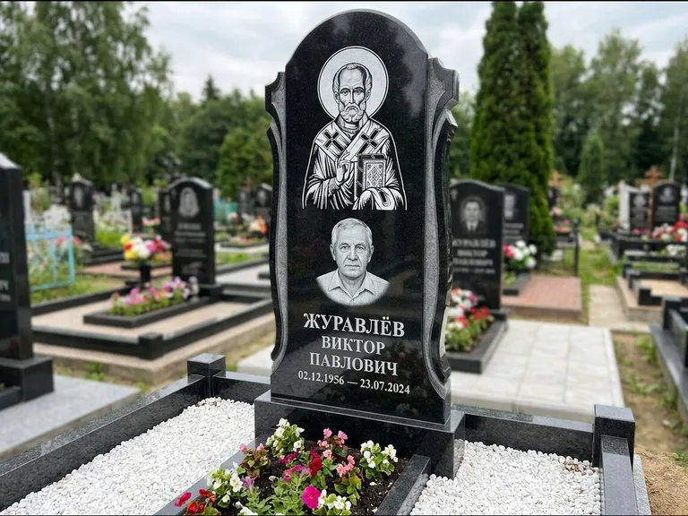 Памятник с гравировкой иконы: стела с образом в полный рост на обороте — гравировка образа Матроны Московской, низкие кусты спиреи в цвету, работа «Лебедева» в Уфе